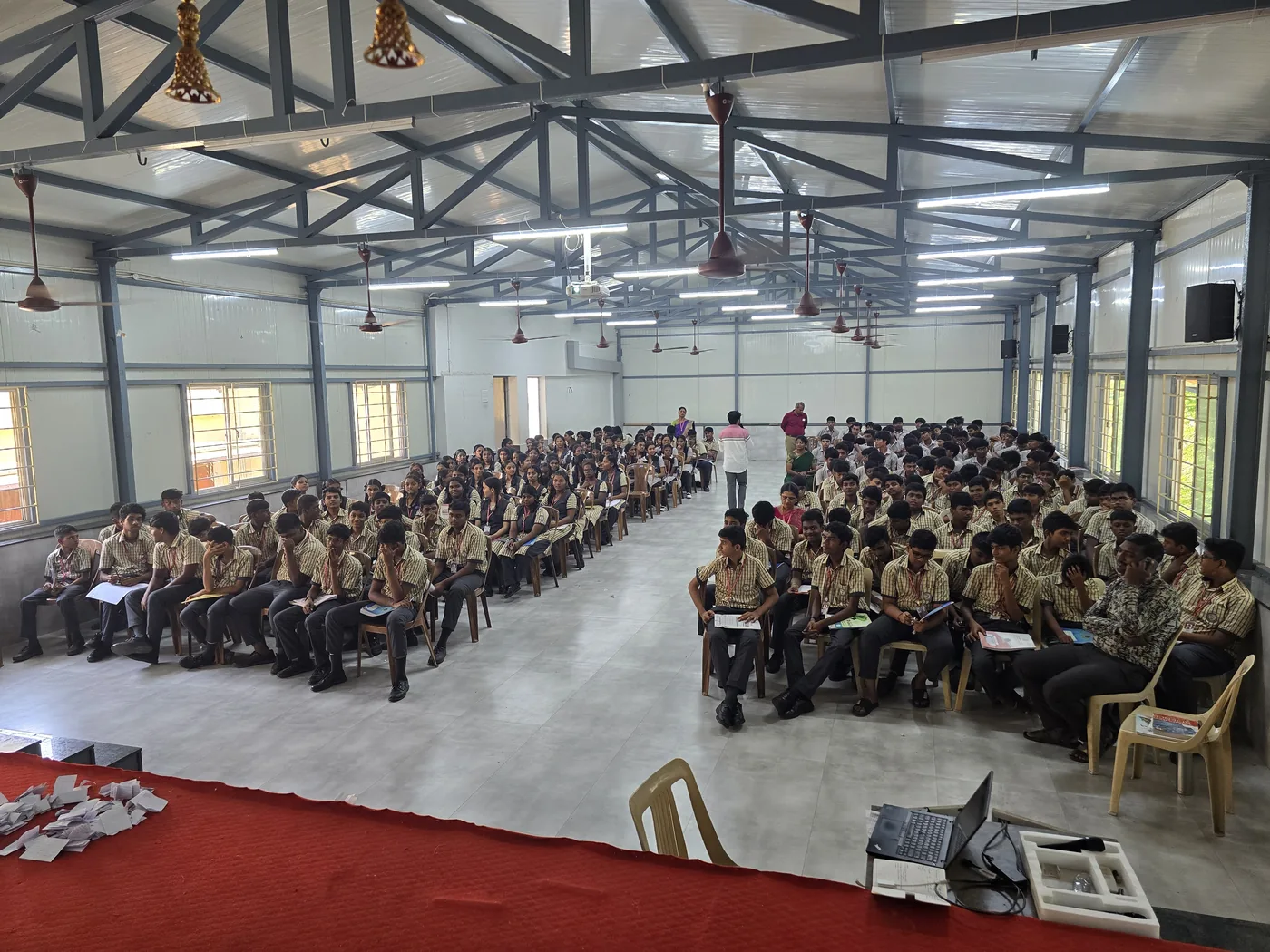 A school hall filled with Class 10, 11 and 12 students in uniform, seated in rows for an OxygenLabs AI workshop.
